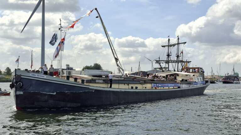 Groot schip met de naam "Abel Tasman" vaart op het water, omringd door mensen aan boord en met een windmolen op de achtergrond.