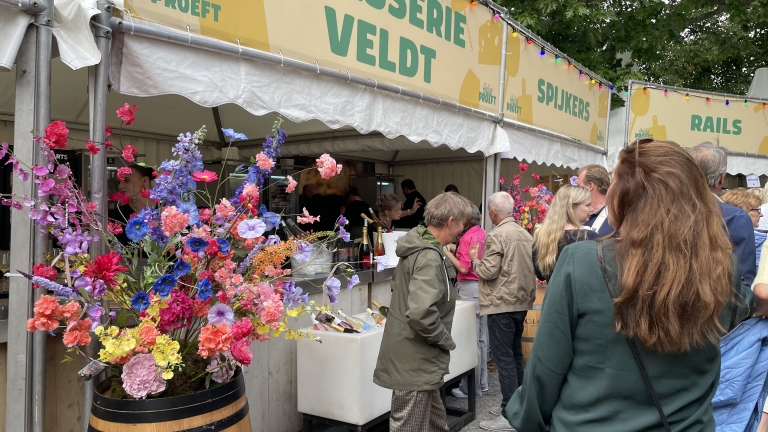 Groep mensen in de rij bij een etenskraam op een buitenfestival, versierd met kleurrijke bloemen en flessen drank in ijs.