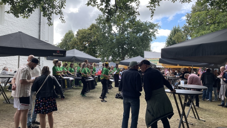Een groep mensen verzameld onder partytenten tijdens een buitenevenement, met muzikanten in groene shirts die spelen op instrumenten. Er zijn tafels en mensen aan het praten terwijl anderen toekijken.