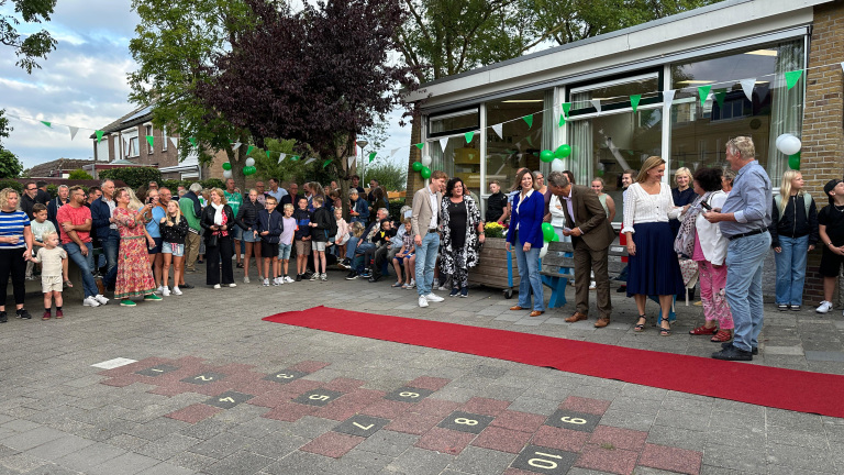 Een groep mensen staat buiten bij een feestelijke gelegenheid met groene decoraties en een rode loper op de grond. Sommigen maken foto's, terwijl anderen kijken of praten. Er zijn volwassenen en kinderen aanwezig bij het evenement.