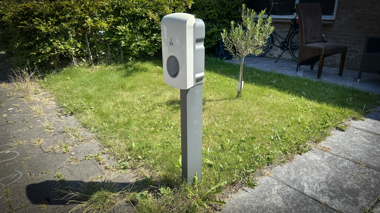 Oplaadpaal voor elektrische auto's op een grasveld naast een geplaveid pad en een kleine boom, met een fiets en stoel in de achtergrond.