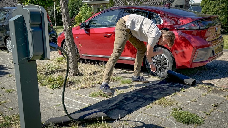 Een persoon legt een rubbermat over een oplaadkabel die is aangesloten op een rode auto, geparkeerd naast het trottoir, om obstakels te vermijden.