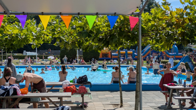 Een drukbezocht openluchtzwembad met veel mensen in zwemkleding, omgeven door groene bomen en kleurrijke vlaggetjes. Op de achtergrond zijn opblaasbare speeltoestellen zichtbaar.