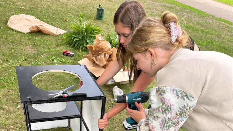 Twee meisjes werken aan een doe-het-zelf project op gras, één gebruikt een boormachine terwijl de ander met een schaartje werkt aan een zwart houten frame met ronde uitsnede.