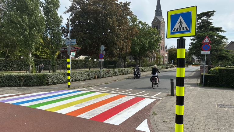 Straatbeeld met een regenboogzebrapad, een fietser en een scooter op de weg, omringd door bomen en een kerk op de achtergrond.