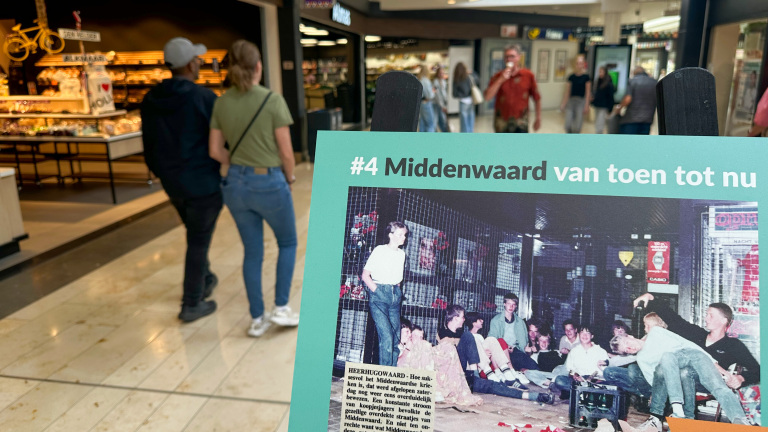 Twee personen lopen door een winkelcentrum; op de voorgrond is een bord te zien met een historische foto en de tekst 'Middenwaard van toen tot nu.'