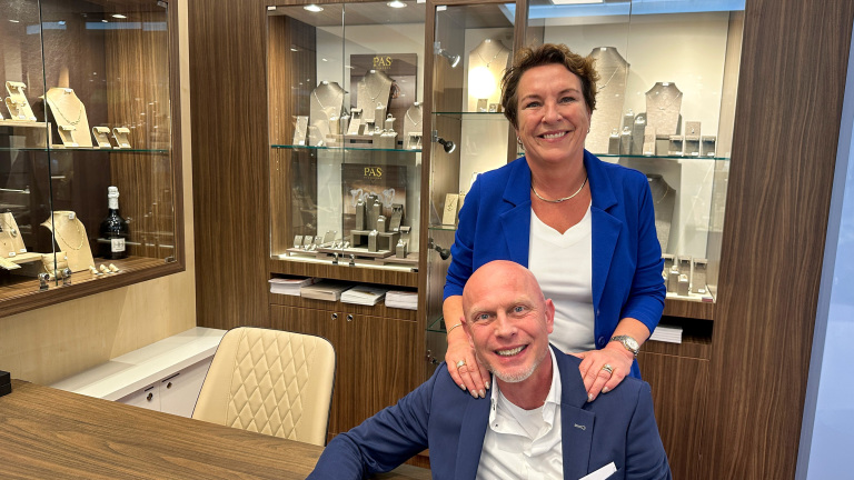 Twee mensen poseren in een juwelierswinkel, omringd door vitrines met sieraden. De vrouw staat achter een zittende man, beide glimlachen en dragen zakelijke kleding.