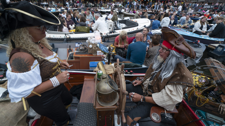 Twee mensen verkleed als piraten zitten op een drukke boot tijdens een festival. De vrouw draagt een zwarte hoed en bruine corset, terwijl de man een rode hoofdband en piratenhoed draagt. Om hen heen zitten mensen op andere boten, terwijl op de achtergrond een drukte van mensen zichtbaar is. Er staat een stuurwiel op de boot en een papegaai zit op de reling.