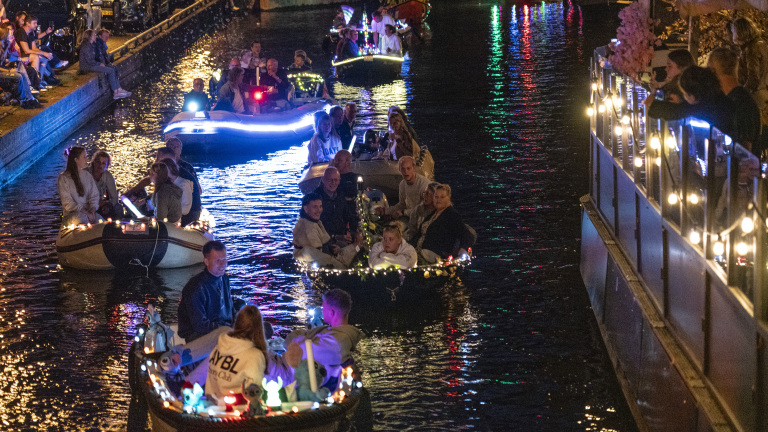 Kleine boten met mensen varen 's avonds over een verlichte gracht, omgeven door toeschouwers langs de kade.
