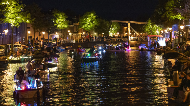 Avondparade van boten versierd met kleurrijke lichten op een verlichte gracht, met feestende mensen langs de waterkant.