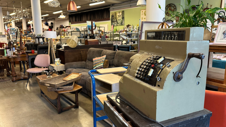 Antieke kassa met een handmatige hendel in een tweedehandswinkel vol met meubels en decoratieve items zoals stoelen, lampen en schilderijen.