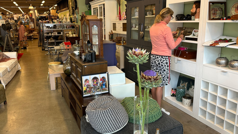 Een vrouw in een kringloopwinkel bekijkt items op een witte plank, omringd door diverse meubels en decoraties.