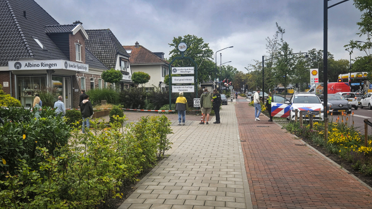 Straatbeeld in een winkelgebied met mensen die langs een juwelier lopen, een politieauto geparkeerd langs de stoep en diverse winkels aan beide zijden. Op de achtergrond zijn verkeersborden en bomen zichtbaar.