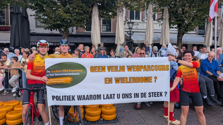 Twee wielrenners en een vrouw in wielerkleding houden een spandoek vast op een plein met toeschouwers. Op het spandoek staat een boodschap die oproept tot veilige wielerfaciliteiten. Op de achtergrond kijken mensen toe bij een evenement.