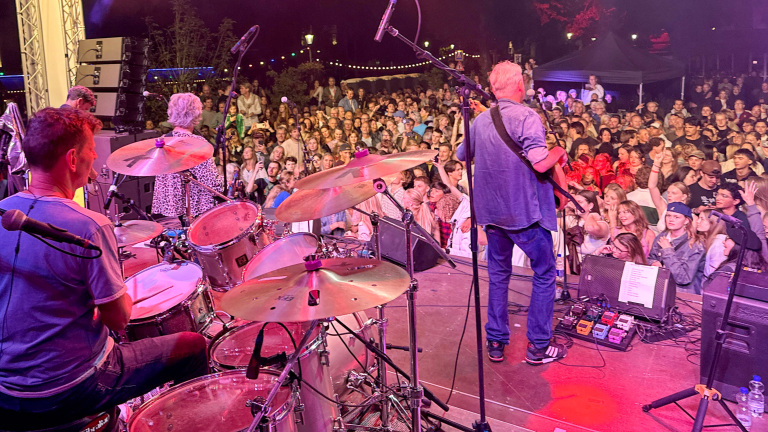 Een band speelt op een podium met een enthousiaste menigte voor hen tijdens een nachtelijk concert. Een drummer, gitarist en andere bandleden zijn zichtbaar, omringd door muziekapparatuur en felgekleurde verlichting.