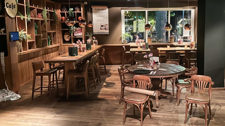 Een gezellig ingerichte eetgelegenheid met houten tafels en stoelen, tafels gedecoreerd met roze bloemen en menukaarten, warme verlichting en planten als decoratie aan de muren.