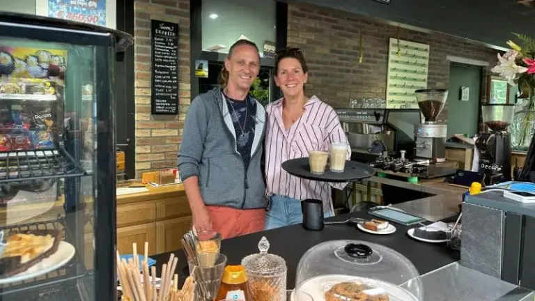 Twee mensen staan glimlachend achter de toonbank van een café, met koffie en gebak zichtbaar op de voorgrond.