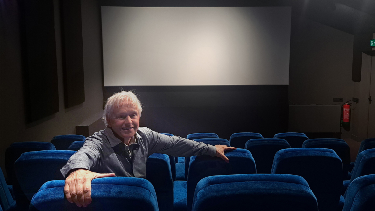 Een oudere man zit lachend in een bijna lege bioscoopzaal met blauwe stoelen, voor een leeg bioscoopscherm.