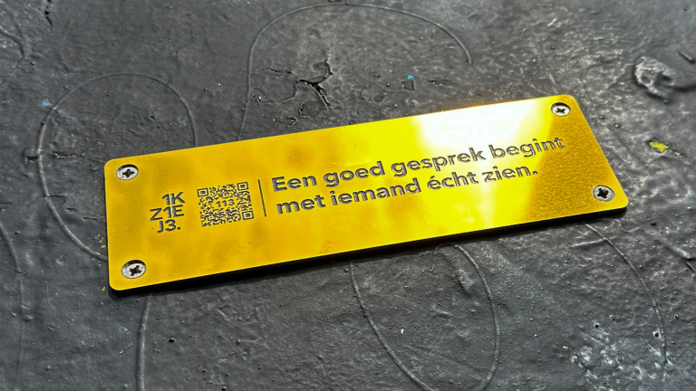 Gouden plaatje met de tekst 'Een goed gesprek begint met iemand écht zien.' en een QR-code erop, bevestigd met schroeven op een donker oppervlak.