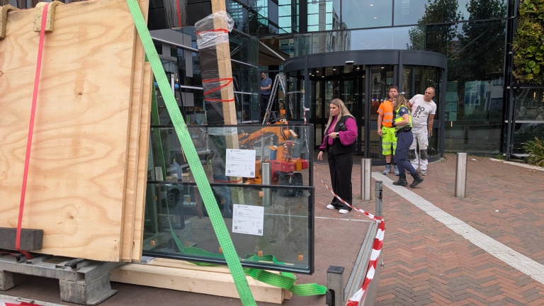 Bouwmaterialen en glazen panelen worden op een houten plaat vervoerd, vastgezet met riemen, voor de glazen entree van een modern gebouw terwijl mensen langs lopen.