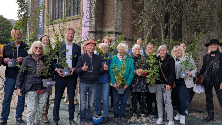 Groep mensen poseert met jonge bomen voor een stenen gebouw met gotische architectuur en een spandoek met de tekst 'Kleine Alm Bezoek'.