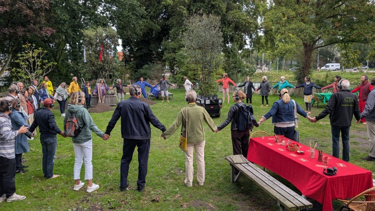 Mensen vormen een cirkel in een bosrijke omgeving, waarbij ze elkaars handen vasthouden en koptelefoons dragen, naast een tafel bedekt met een rood tafelkleed.