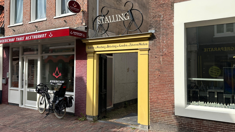 Een Thais restaurant genaamd Khunchai met een fiets geparkeerd voor de deur en een gele doorgang naar een fietsstalling aan de rechterkant van een straat in een stedelijke omgeving.