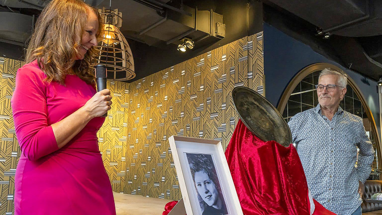 Een vrouw in een roze jurk spreekt in een microfoon terwijl ze naar een ingelijste zwart-witfoto kijkt, naast een man in een blauw shirt en een frisdrankblikje op een rood fluwelen doek in een kamer met geometrisch behang.