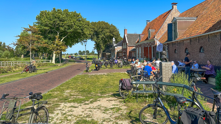 Mensen zitten buiten een bakstenen gebouw met oranje dak pannen, omringd door geparkeerde fietsen langs een smalle geplaveide weg op een zonnige dag in een landelijke omgeving.