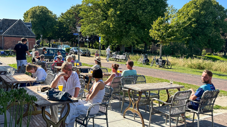 Mensen genieten van drankjes op een zonnig terras, omgeven door bomen en een rustige straat met geparkeerde fietsen.