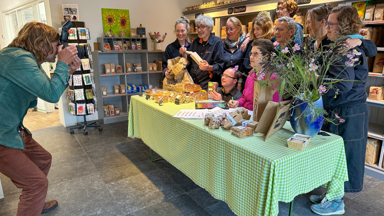 Een fotograaf maakt een foto van een groep mensen die poseren achter een tafel met bakwaren en planten, in een winkelomgeving met stellingen vol producten op de achtergrond.