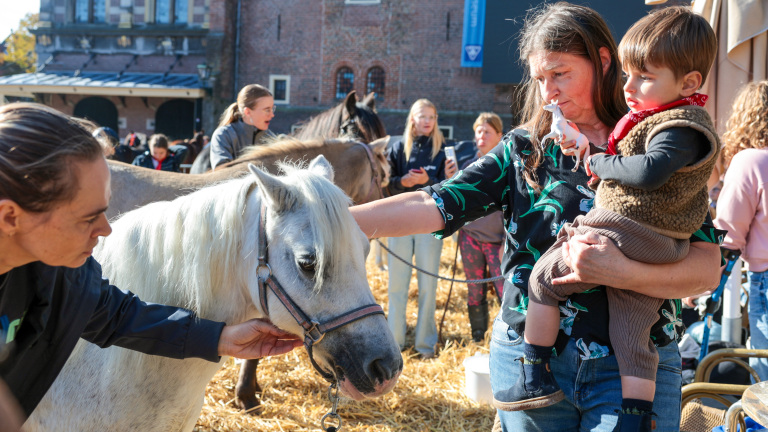Een vrouw houdt een kind vast, die een speelgoedpaardje vasthoudt, terwijl een andere vrouw een wit paard aait in een omheinde buitenruimte met stro, omgeven door andere mensen en paarden.