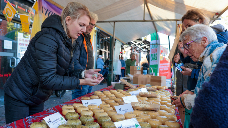 Mensen op een markt staan bij een kraampje met verschillende soorten kaas op een tafellaken met rode patronen.