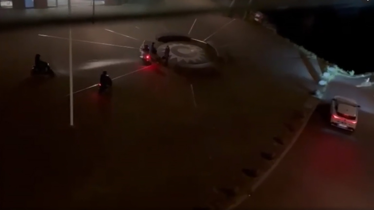 Nachtbeeld van een verlichte rotonde waar motoren en een auto rijden onder natte omstandigheden.