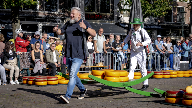 Een man in een zwart shirt en een man in een wit kostuum en groene hoed dragen samen een kaasbrancard tijdens een evenement op een plein, terwijl een menigte toekijkt.