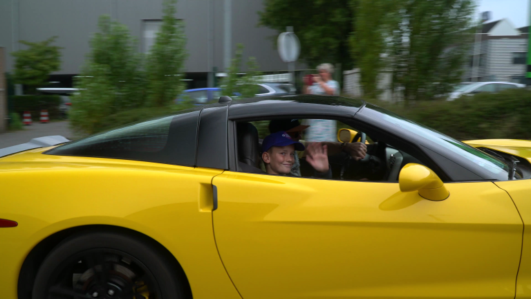 Een jongen met een blauwe pet zwaait vanuit een gele sportauto met zwarte velgen, terwijl de auto langs een groene omgeving rijdt.