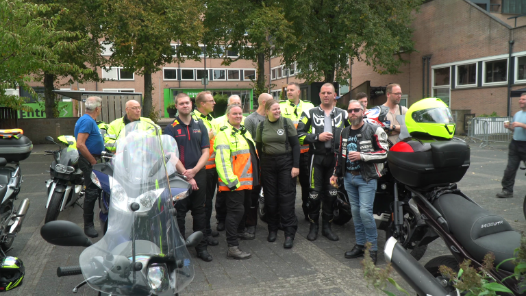 Een groepje motorrijders in kleurrijke veiligheidskleding staat samen met hun motorfietsen voor een gebouw.