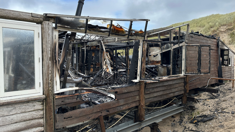Uitgebrand houten gebouw in de duinen, met zwartgeblakerde resten en gebroken ramen, onder een bewolkte hemel.