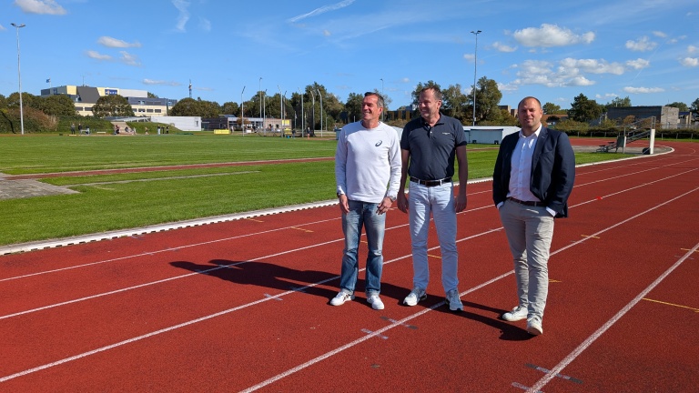 Drie mannen staan op een atletiekbaan met een groene grasmat en een gebouw in de achtergrond op een zonnige dag.