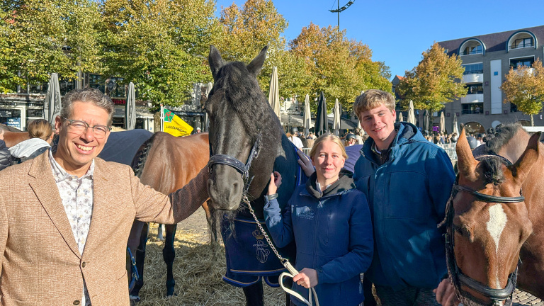 Groep mensen poseert met paarden op een plein omringd door bomen en gebouwen op een zonnige dag.
