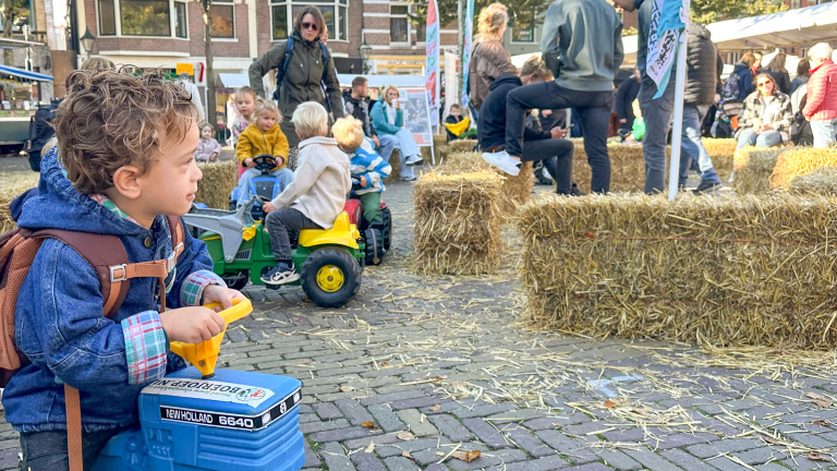 Een jong kind zit op een blauwe speelgoedtractor tussen hooibalen op een buitenmarkt waar meerdere kinderen spelen en volwassenen toekijken.