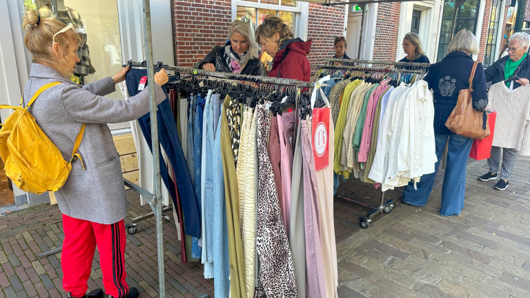 Mensen bladeren door kledingrekken met diverse broeken en blouses op een trottoir buiten een winkel.