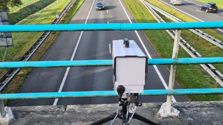 Snelheidscamera op statief boven een viaduct dat uitkijkt op een snelweg met passerende auto's.
