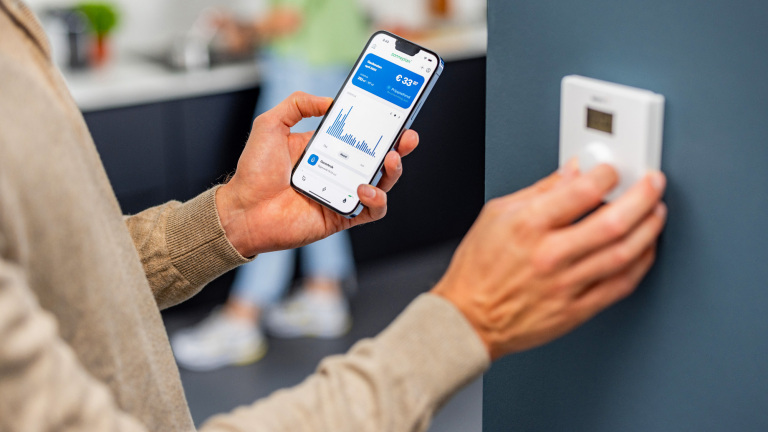 Persoon bedient een thermostaat op de muur terwijl hij een smartphone vasthoudt met een energiemanagement-app die grafieken en een prijsweergave toont.