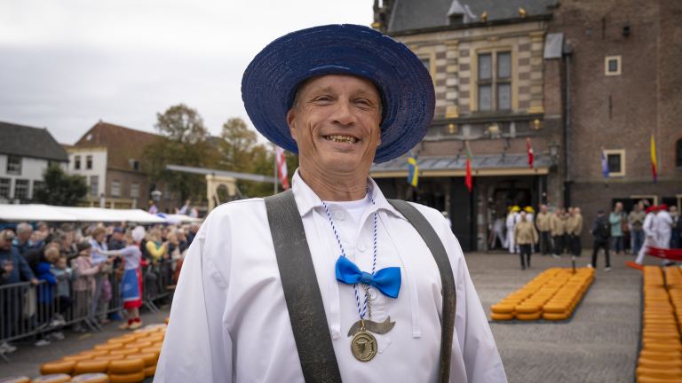 Man in traditional Dutch attire with a blue hat and bow tie, smiling at a cheese market scene, with rows of cheese wheels and a crowd in the background.