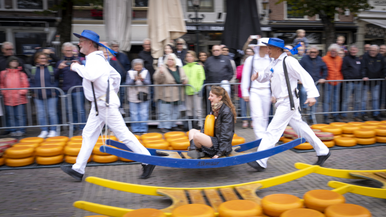 Twee kaasdragers in witte uniformen en blauwe hoeden rennen met een oplegger waarop een vrouw zit, omringd door kazen, tijdens een evenement voor toeschouwers.