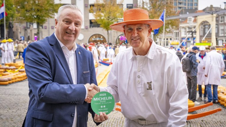 Twee mannen schudden elkaar de hand op een druk plein vol mensen en kaasrondeals, waarbij een man in een wit pak en oranje hoed een Michelin-bordje voor 2025 vasthoudt.