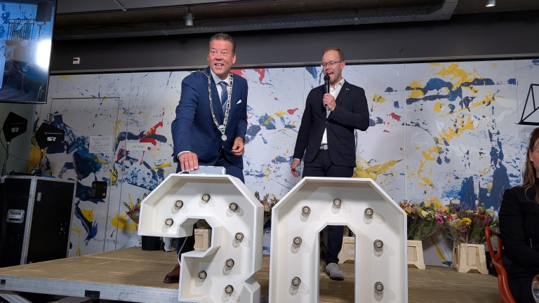 Twee mannen in pak staan op een podium met een feestelijke achtergrond; een van hen houdt een microfoon vast terwijl de ander lacht naast grote verlichte cijfers "20". Op de achtergrond staan bloemen.