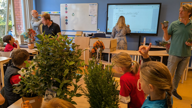 Kinderen in een klaslokaal zitten bij tafels met potplanten, terwijl een vrouw op een digibord schrijft en een man vooraan potplanten vasthoudt.