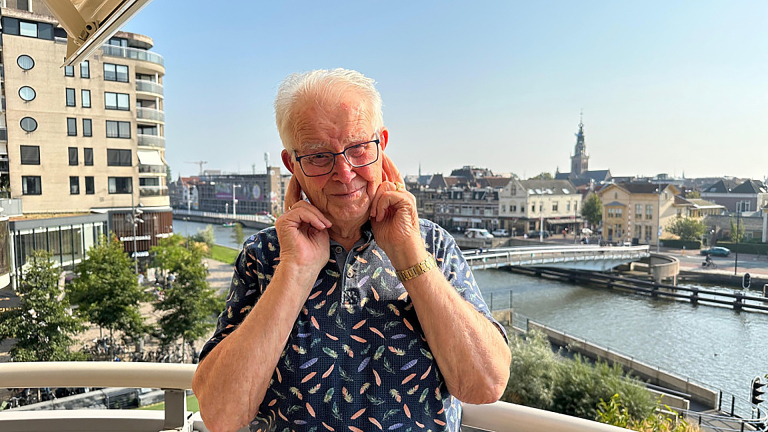 Een oudere man met een bril staat op een balkon met de stad en een kanaal op de achtergrond, terwijl hij zijn vingers in zijn oren stopt.